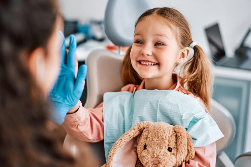 At The Doctor's Appointment. A Candid Emotional Photo Of A Child Sitting In A Dental Chair, Holding A Toy Rabbit And Cheerfully Giving A High Five To The Nurse.