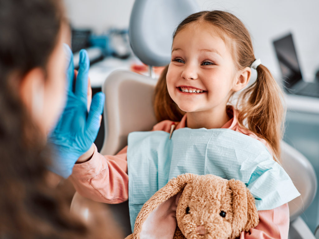 At The Doctor's Appointment. A Candid Emotional Photo Of A Child Sitting In A Dental Chair, Holding A Toy Rabbit And Cheerfully Giving A High Five To The Nurse.